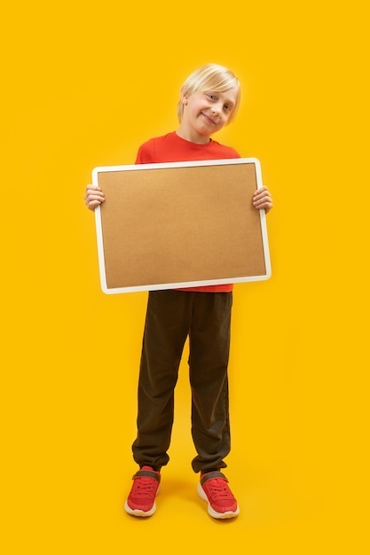 Cornice verticale fulllength ritratto di ragazzo biondo con tavola corticale nelle sue mani sfondo giallo Posto per pubblicizzare Copia spazio