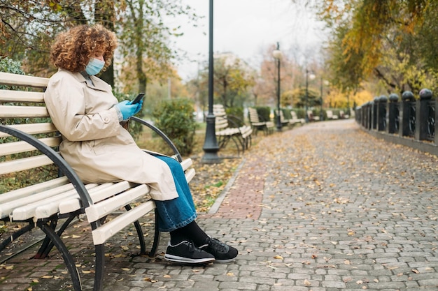 Corona all'aperto Quarantena attiva Passeggiata nel parco Capelli ricci concentrati Donna in sovrappeso in maschera chirurgica al di fuori del testo del telefono digitando sulla panca nello spazio di copia della vista del parco autunnale