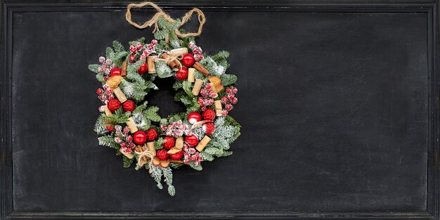 Corona di Natale fatta di rami di abete, mele secche, cannella, bacche rosse, tappi di bottiglia, palline rosse appese su una tavola di gesso nera.