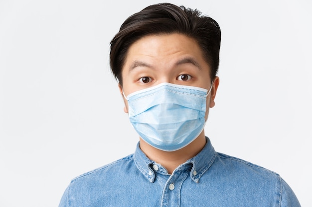 Coronavirus, distanza sociale e concetto di stile di vita. Uomo asiatico sorpreso in maschera medica che alza le sopracciglia in soggezione, in piedi stupito su sfondo bianco, usando misure protettive durante il covid-19.