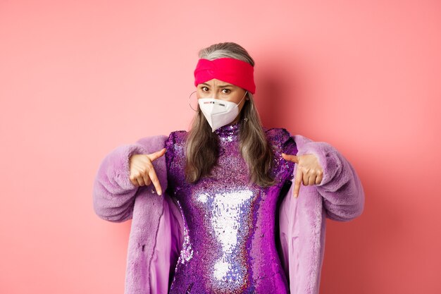 Coronavirus e concetto di acquisto. Elegante donna anziana asiatica in abiti viola e maschera facciale, puntando le dita verso il basso, mostrando promo, sfondo rosa.