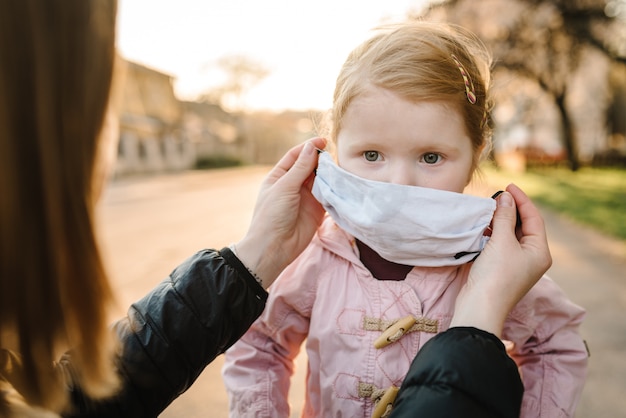 Coronavirus e concetto di inquinamento atmosferico. Le maschere d'uso della madre e della bambina camminano sulla via. La mamma corregge la maschera del bambino. Sintomi del virus pandemico. Famiglia con bambino all'aperto. Protezione delle malattie.