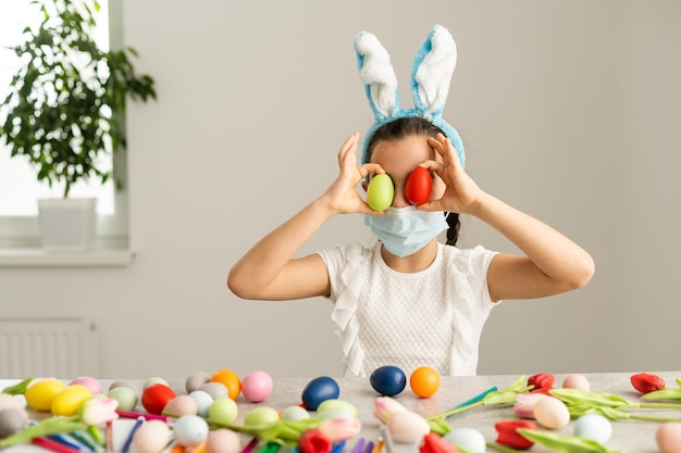 Coronavirus e vacanze di Pasqua. Bambina sveglia in una maschera medica protettiva con orecchie da coniglio con uova di Pasqua e fiori primaverili.