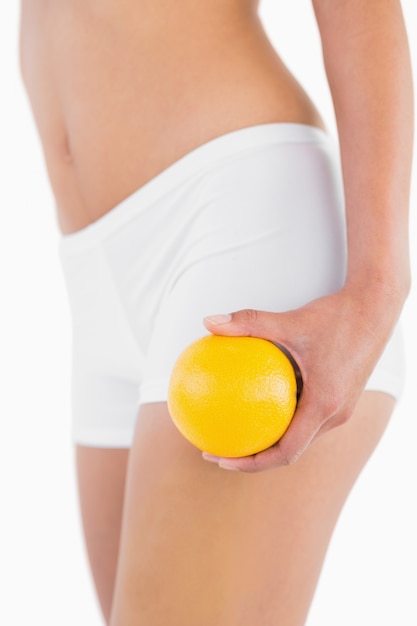 Corpo femminile snello che tiene arancia sulla sua gamba su sfondo bianco