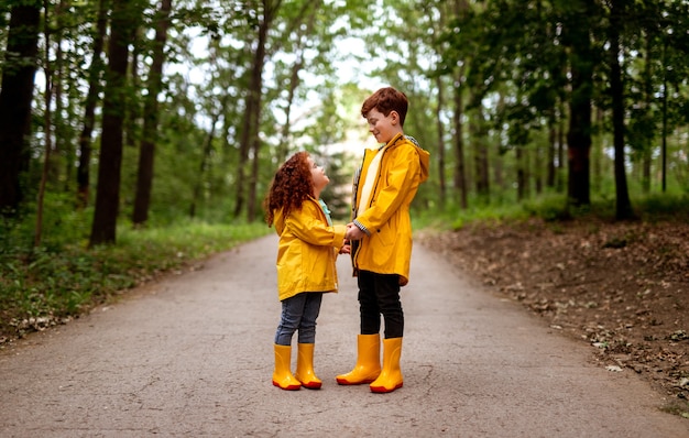 Corpo pieno di bambina sorridente e fratello maggiore con i capelli rossi vestiti con impermeabili gialli brillanti e stivali di gomma che si tengono per mano e si guardano, mentre si trovano in una passerella nella foresta verde