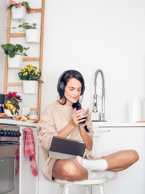 Corpo pieno di donna ispanica concentrata con le cuffie seduta con una tazza di caffè e un tablet in cucina