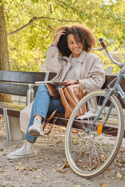 Corpo pieno di giovane signora etnica positiva ed elegante che riposa con il libro sulla panca di legno vicino alla bicicletta nel parco e tocca i capelli