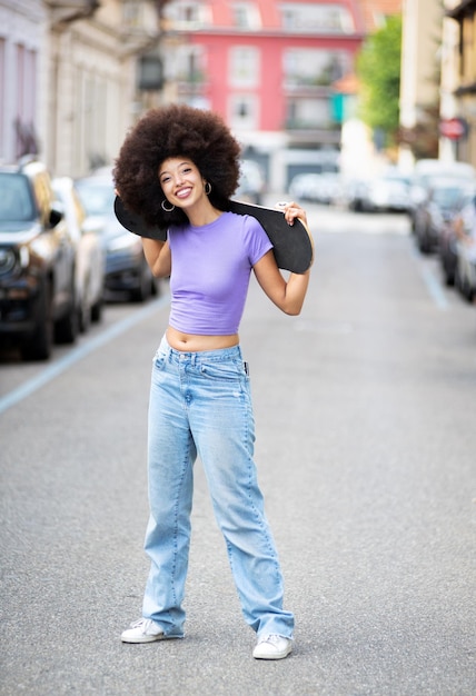 Corpo pieno di una giovane donna afroamericana positiva con i capelli ricci che posa con lo skateboard in una strada della città