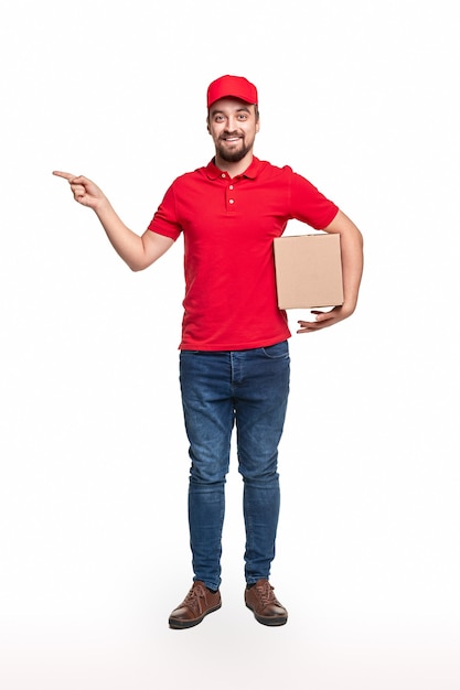 Corpo pieno di uomo di consegna positivo in camicia rossa e cappuccio che trasportano la scatola di cartone e che punta allo spazio vuoto