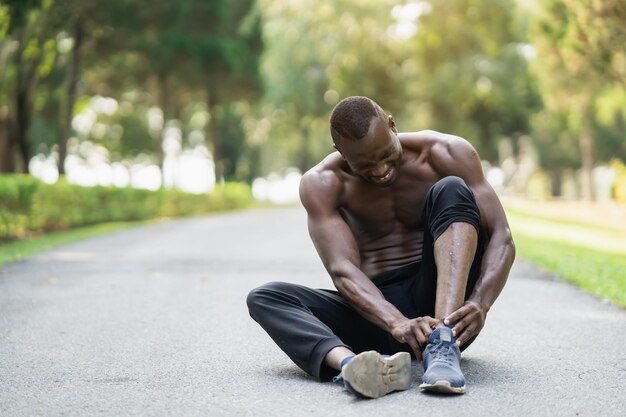 Corridore africano uomo seduto sulla pista da jogging e sensazione di dolore alla caviglia, copia spazio. Concetto di infortunio sportivo