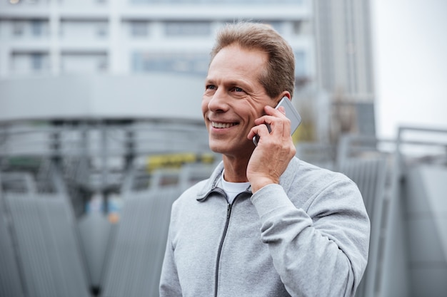 Corridore sorridente in abiti sportivi grigi che parla al telefono per strada. Vista laterale