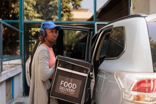 Corriere afroamericano che consegna cibo per ufficio in auto ritratto, in attesa di clienti all'aperto. Addetto al servizio di consegna che estrae lo zaino dal veicolo, guardando la fotocamera