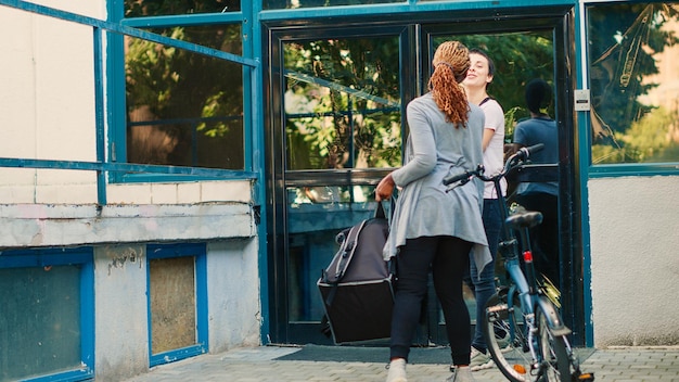 Corriere afroamericano che dà il pasto del sacchetto di carta al cliente femminile, consegnando il pacchetto di cibo del ristorante in bicicletta. Donna che riceve un fast food da asporto dal corriere, seduto all'ingresso dell'edificio.