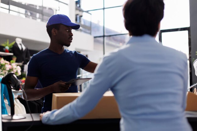 Corriere afroamericano che spiega i dettagli della spedizione al lavoratore, prendendo le scatole di cartone dal bancone del centro commerciale. Manager che prepara i pacchi per la consegna, lavora in una boutique moderna