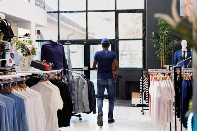 Corriere afroamericano che trasporta scatole di cartone, lasciando il negozio di abbigliamento. Uomo che indossa l'uniforme di consegna, si prepara a spedire i pacchi dei clienti nella moderna boutique. Concetto di moda