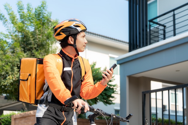 Corriere asiatico dell'uomo che controlla l'indirizzo del cliente in mappa in telefono sulla bicicletta che consegna alimento nelle vie della città con una consegna calda dell'alimento da take away e ristoranti a casa.