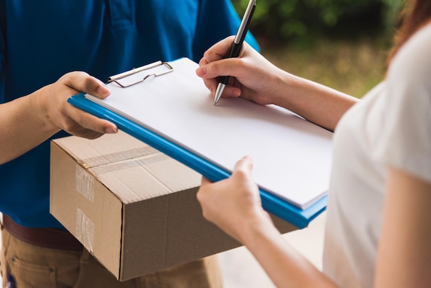 Corriere asiatico giovane fattorino che fa acquisti online con pacco postale in maschera protettiva uniforme coronavirus che fa servizio cliente donna utilizzando la firma della penna sul libro cartaceo a casa, COVID-19