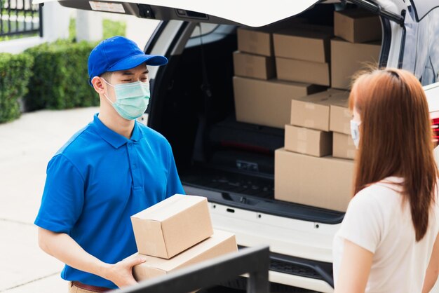 Corriere asiatico giovane fattorino che fa acquisti online consegna pacco postale lui maschera protettiva e cliente donna di servizio che riceve inscatolato davanti alla porta di casa, sotto il coprifuoco pandemia coronavirus COVID-19