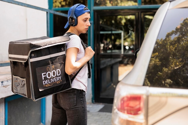 Corriere con borsa termica che consegna l'ordine alimentare in auto, portando lo zaino con il pranzo. Servizio di consegna pasti in ufficio giovane dipendente che ascolta musica in cuffia, vista laterale