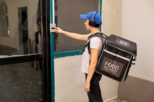 Corriere della donna del servizio di consegna del cibo che suona il campanello dell'edificio, in attesa del cliente all'aperto. Ragazza in cuffia con zaino termico in piedi davanti alla porta, che consegna il pranzo da asporto