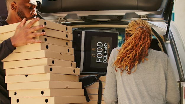 Corriere femminile afroamericano che prende un enorme mucchio di scatole per pizza dal bagagliaio del veicolo, consegnando un grande ordine di pasti da pizzeria ai clienti alla porta d'ingresso. Usando l'auto per consegnare una pila di fastfood.