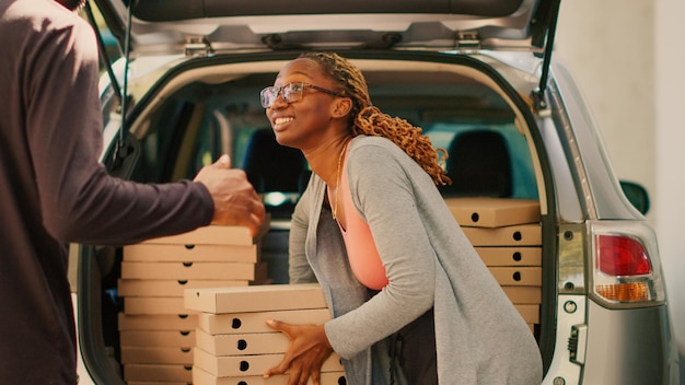 Corriere femminile che consegna scatole per pizza impilate dal bagagliaio dell'auto, prendendo un enorme ordine di pizzerie in pile fuori dal veicolo. Addetto alle consegne da asporto che consegna pacchi ai clienti in piedi. Tiro a mano libera.