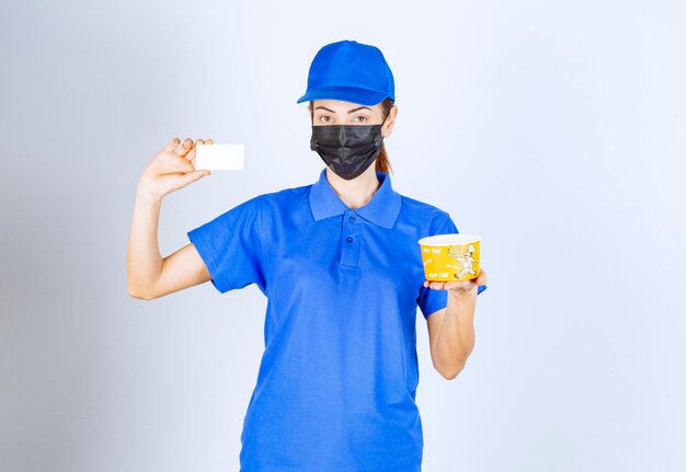 Corriere femminile del ristorante in uniforme blu e maschera facciale che consegna un cibo da asporto e presenta il suo biglietto da visita.