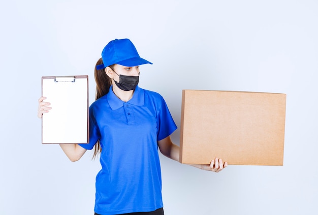 Corriere femminile in maschera e uniforme blu che tiene un grosso pacco di cartone e presenta la lista di controllo per la firma