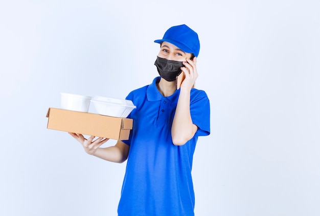 Corriere femminile in maschera e uniforme blu che tiene una scatola di cartone, pacchi da asporto e parla al telefono.