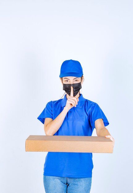 Corriere femminile in uniforme blu e maschera facciale che tiene una scatola di cartone e chiede silenzio.