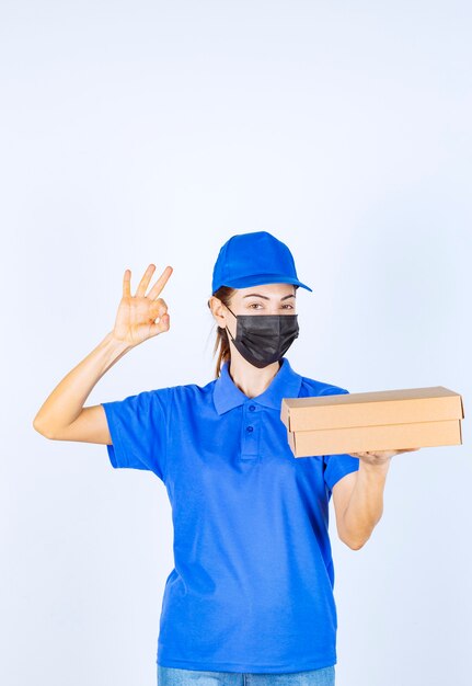 Corriere femminile in uniforme blu e maschera facciale che tiene una scatola di cartone e mostra il segno della mano di godimento.