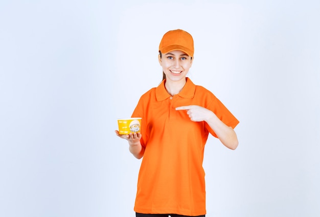 Corriere femminile in uniforme gialla che tiene una tazza di tagliatelle da asporto e indicandola.