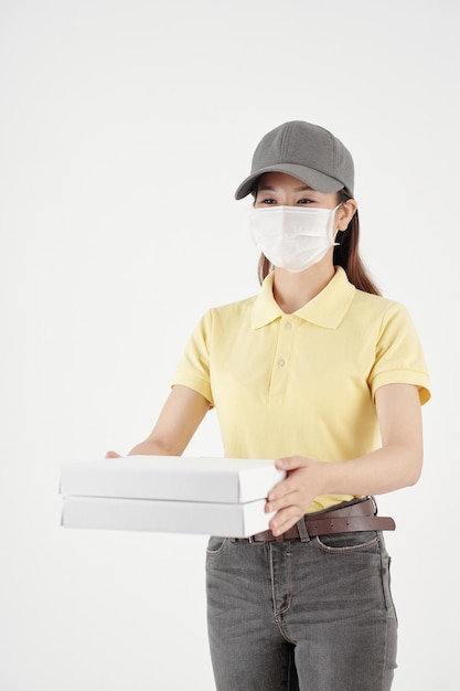 Corriere femminile sorridente in maschera medica che dà scatole con pizza calda isolata su bianco