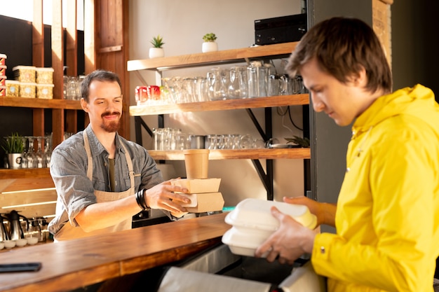 Corriere in giacca gialla che mette le scatole per il pranzo nella borsa termica mentre il cameriere gli passa una pila di scatole di cibo nel caffè