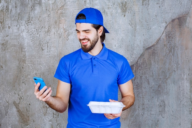 Corriere in uniforme blu che tiene una scatola di plastica da asporto e prende nuovi ordini via telefono