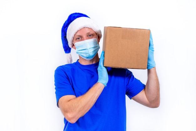 Corriere in uniforme blu, maschera medica protettiva, guanti e cappello di Babbo Natale tiene la confezione regalo