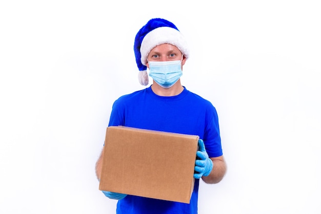 Corriere in uniforme blu, maschera medica protettiva, guanti e cappello di Babbo Natale tiene la confezione regalo