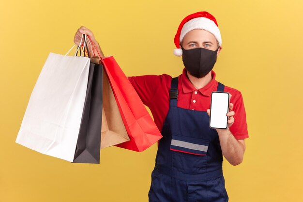 Corriere maschile in maschera medica protettiva e cappello rosso di Babbo Natale con borse della spesa e smartphone servizi di quarantena per app di ordine e consegna facili Colpo in studio al coperto isolato su sfondo giallo