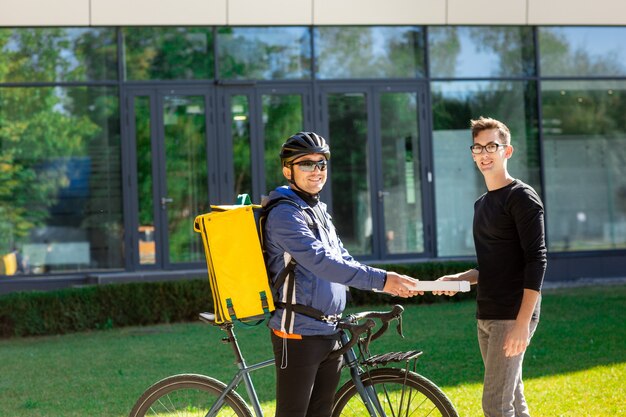 Corriere maschio con la bicicletta e la borsa termica che danno il contenitore di pizza al cliente sulla via vicino all'ufficio