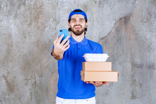 Corriere maschio in uniforme blu che trasporta scatole di cartone e plastica e prende nuovi ordini tramite smartphone.