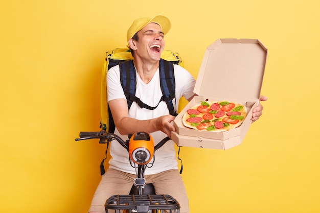 Corriere ottimista divertente uomo sulla bici che tiene scatola di cartone per pizza e ridendo esprimendo emozioni positive consegnando cibo per ciclo indossando maglietta bianca e cappuccio isolato su sfondo giallo