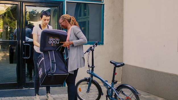 Corriere per la consegna di cibo che dà un pasto in un sacchetto di carta a una donna, che consegna cibo al ristorante in bicicletta. Andare in bicicletta e portare lo zaino per consegnare il pacchetto fast food da asporto all'ingresso dell'edificio.