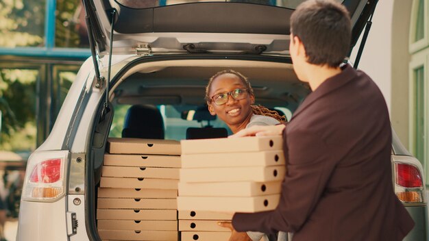 Corriere pizzeria donna che consegna un grosso ordine di fastfood dal bagagliaio del veicolo, dando pile di scatole per pizza alla cliente femminile in piedi all'ingresso dell'edificio. Addetto alla consegna di cibo con pacchi pasto.