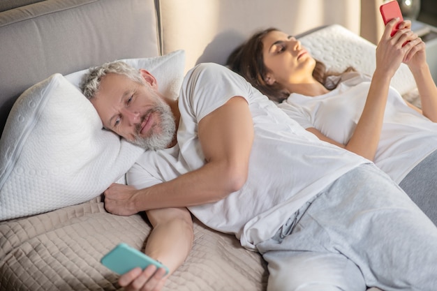 Corrispondenza personale. Giovane donna e guardando lontano sorridente uomo adulto sdraiato con gli smartphone a casa in camera da letto