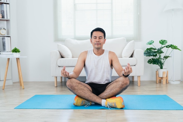 Corso di formazione online Felice uomo maturo asiatico in abbigliamento sportivo che fa stretching per le gambe sul tappetino yoga e medita nell'interno del soggiorno e sorride Felice allenamento sportivo da parte sua slef Sport a casa