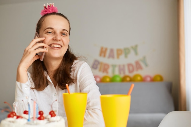 Cortometraggio interno di una giovane donna felice sorridente che indossa una camicia bianca che celebra il compleanno seduto al tavolo con una torta e un telefono che ha soddisfatto l'espressione facciale