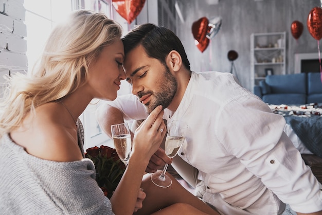 È così bello stare insieme! Bella giovane coppia che beve champagne e sorride mentre è seduta faccia a faccia in camera da letto