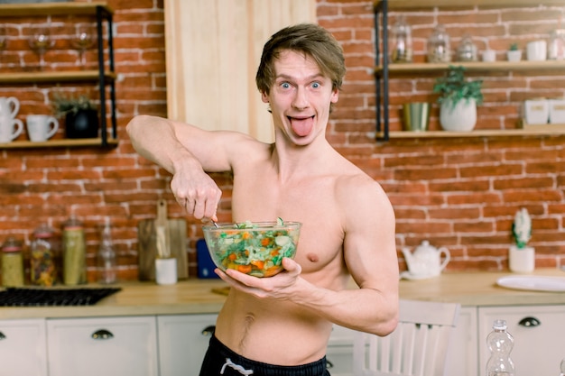 È così delizioso Il giovane felice casuale che prepara l'insalata a casa nella cucina del sottotetto e esamina la macchina fotografica e gli spettacoli è lingua