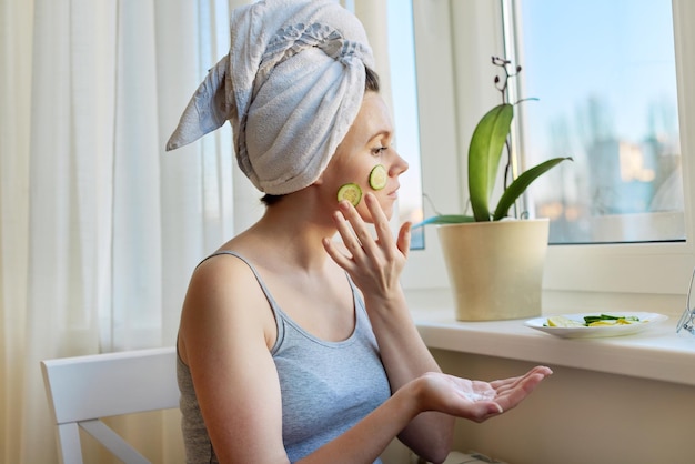 Cosmetici naturali fatti in casa giovane donna con asciugamano da bagno sulla testa applicando crema e fette di cetriolo sul viso seduto vicino alla finestra di casa