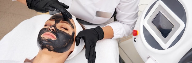 Cosmetologo applicando maschera nera sul viso di una bella donna per la buccia del carbonio